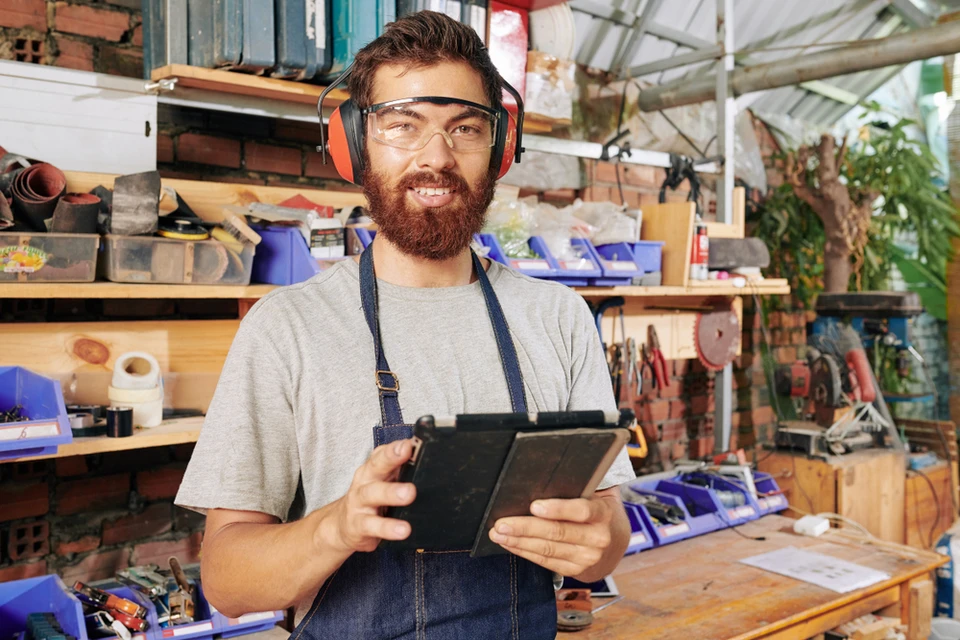 Atelier artisanal avec homme barbu portant tablier bleu, lunettes de sécurité et casque antibruit, tenant une tablette numérique